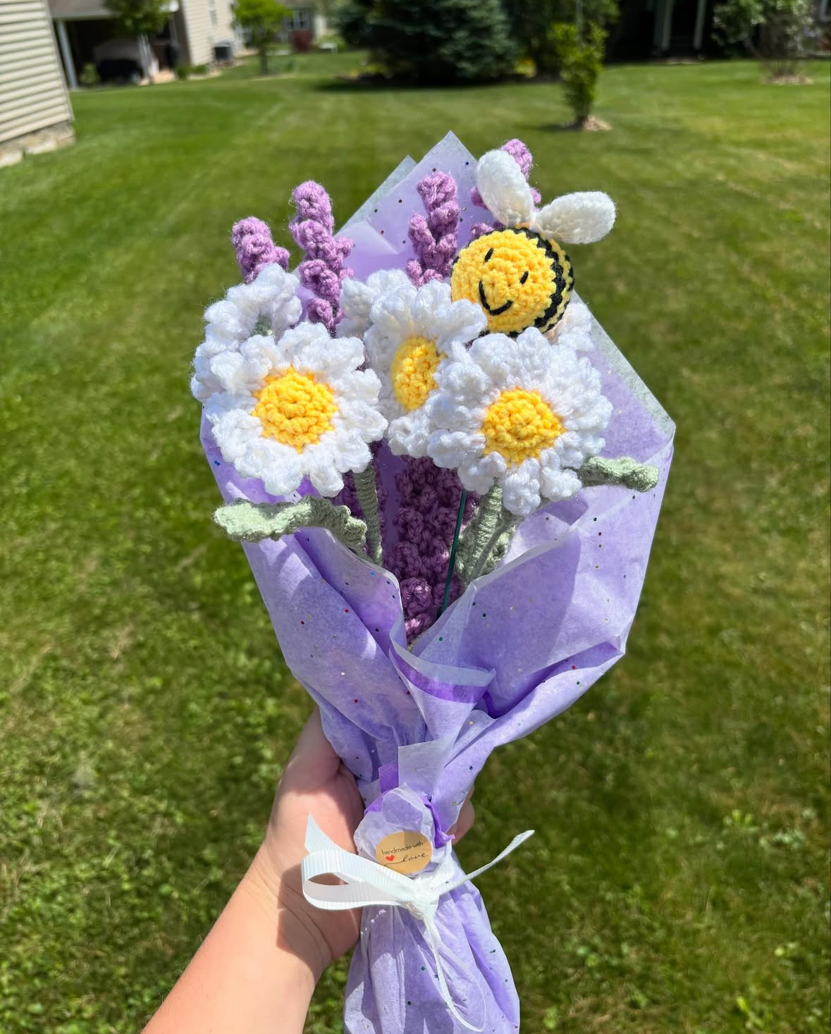 A hand-crafted crochet bouquet featuring white daisies with yellow centers, purple lavender sprigs, and a small amigurumi honeybee, wrapped in light purple paper with a white ribbon.